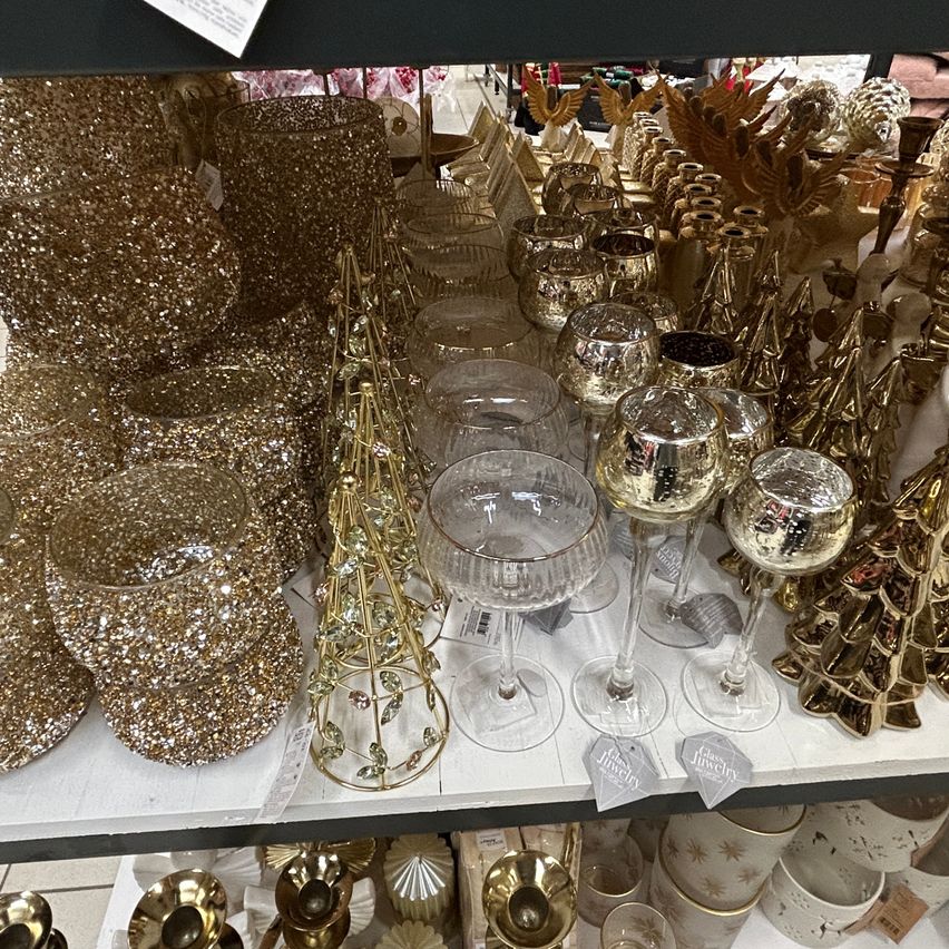 Ein Ladenregal zeigt eine Sammlung von goldplattierten Weingläsern, glitzernden Vasen und verschiedenen dekorativen Gegenständen, einschließlich Glas- und Metallornamenten. Die Umgebung deutet auf eine festliche oder weihnachtlich-thematische Anordnung hin.