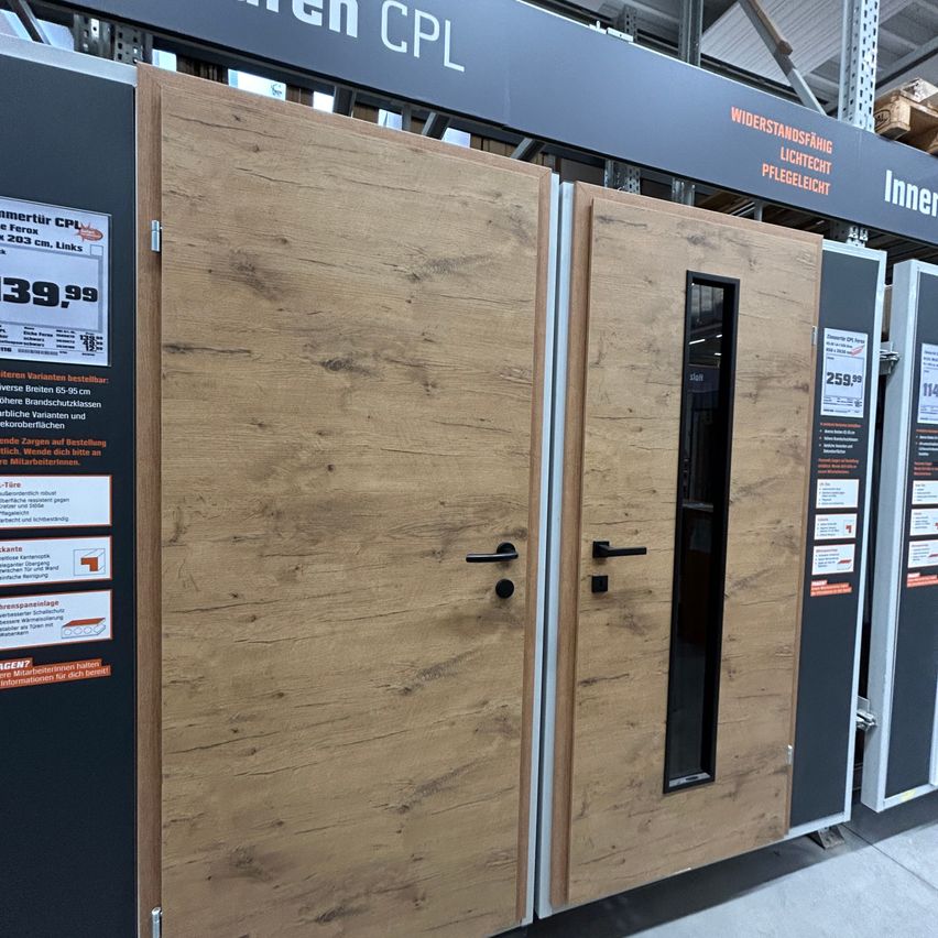 Eine Holztür mit einem Glasfenster und einem schwarzen Griff wird in einem Ausstellungsraum präsentiert. Auf beiden Seiten befinden sich zwei weitere Türen. Hinter der Tür befindet sich eine schwarze Wand mit Text.
