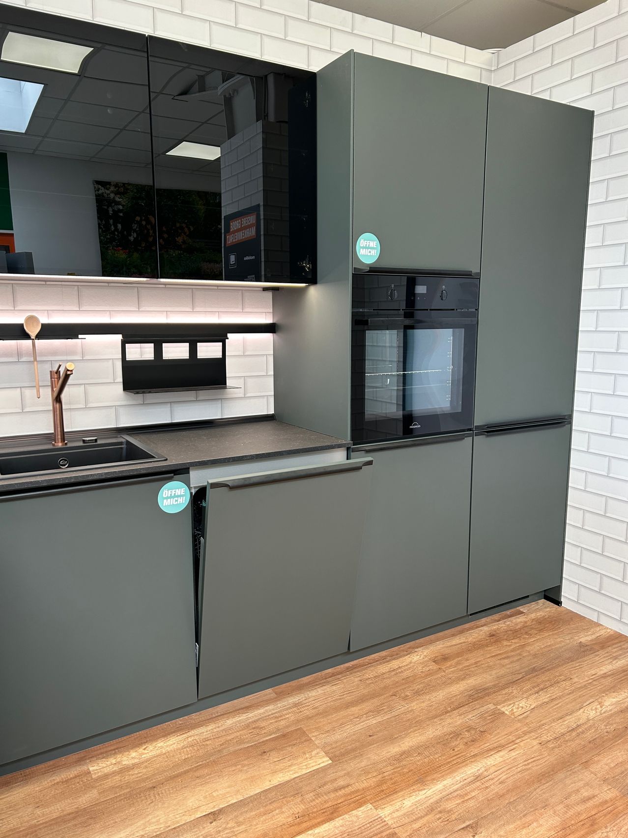 Eine moderne Küche mit schwarzem Backofen, Holzboden und grauen Schränken. Die Arbeitsplatte ist mit einem Waschbecken und einem Wasserhahn ausgestattet. Es gibt auch Aufkleber an den Schränken.