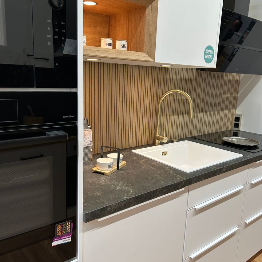 Eine moderne Küche mit einem weißen Waschbecken, goldenem Wasserhahn und Granitarbeitsplatte. Die Rückwand besteht aus vertikalen Holzlatten. Links sind Mikrowelle, Ofen und Kühlschrank.