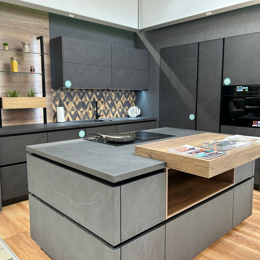 Eine moderne Küche mit dunkelgrauen Schränken, Holzarbeitsplatten und einem schwarzen Herd oben. Die Wand ist grau mit geometrischen Mustern. Regale mit Pflanzen und Gläsern befinden sich links.