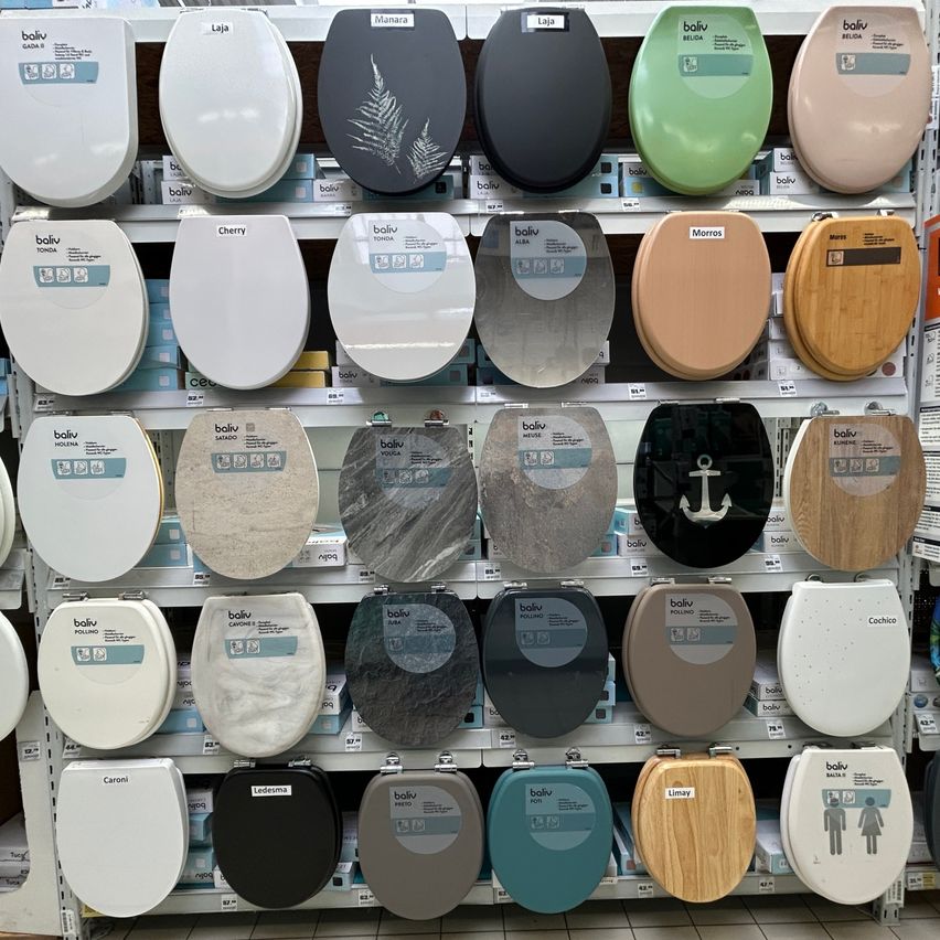 Ein Geschäft zeigt eine Auswahl an Toilettensitzen in verschiedenen Farben und Designs, einschließlich weiß, grün, grau und Holz. Die Sitze sind in Regalen aufgestellt, einige mit Etiketten und andere mit dekorativen Designs.