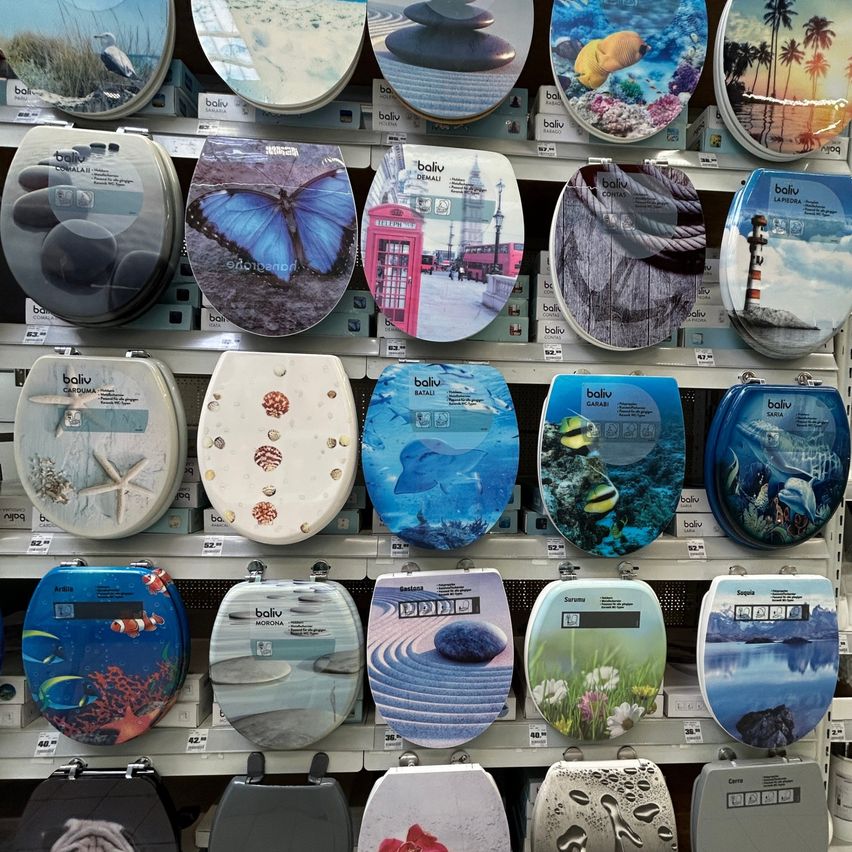 Eine Vielzahl von Toilettendeckeln mit verschiedenen Designs ist auf einem Regal ausgestellt, darunter solche mit Schmetterling, Korallenriff und Londoner Telefonzelle.