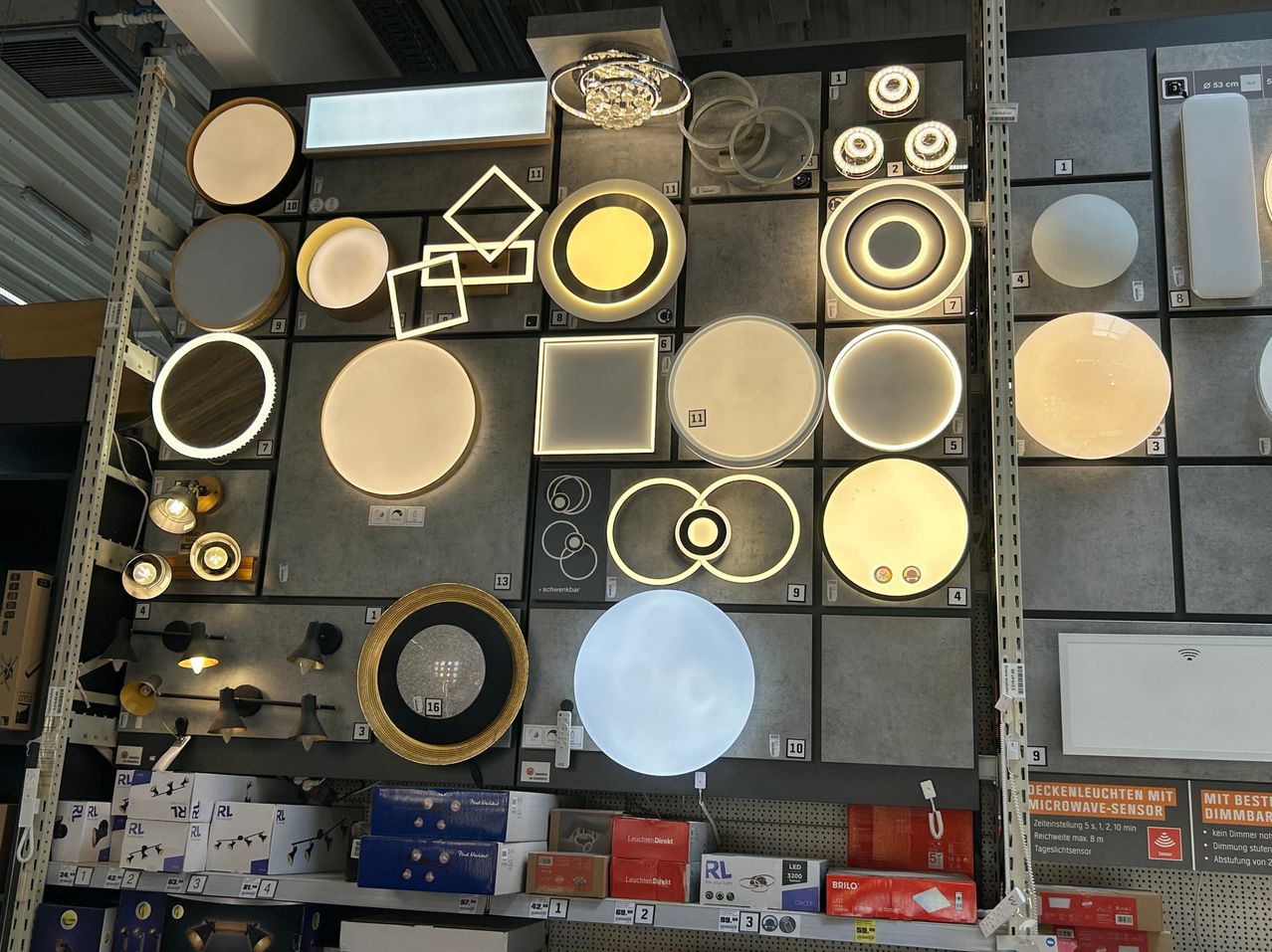 Eine Auswahl an Lampen ist an einer Wand ausgestellt. Einige Lampen sind beleuchtet und andere nicht. Die Lampen sind rund und quadratisch, mit verschiedenen Farben und Designs.