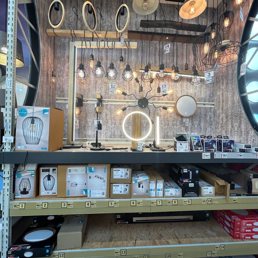 Ein Geschäft zeigt verschiedene Beleuchtungskörper und Lampen auf Holzregalen. Verschiedene Stile werden präsentiert, darunter Hängelampen, Tischleuchten und Deckenleuchten. Kartons mit Produktdetails sind ordentlich unter den Leuchten angeordnet.