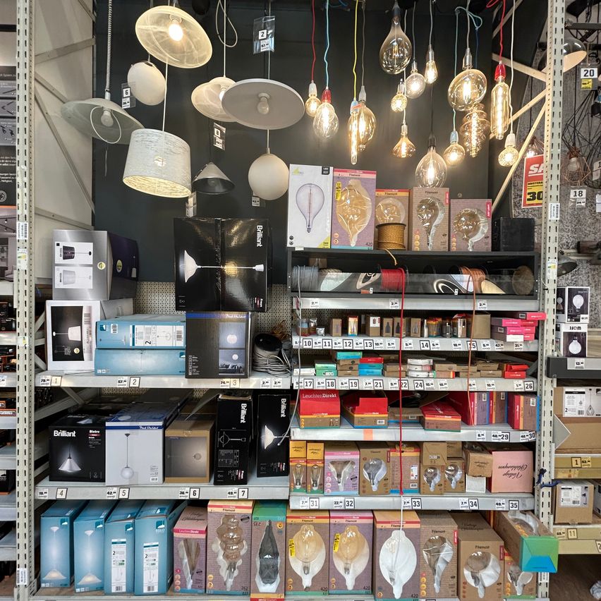 Ein Geschäft mit einer Vielzahl von Beleuchtungsprodukten zur Schau, einschließlich Lampen, Glühbirnen und Zubehör. Die Regale sind mit Kartons und Produkten gefüllt, und einige Artikel hängen an der Decke.