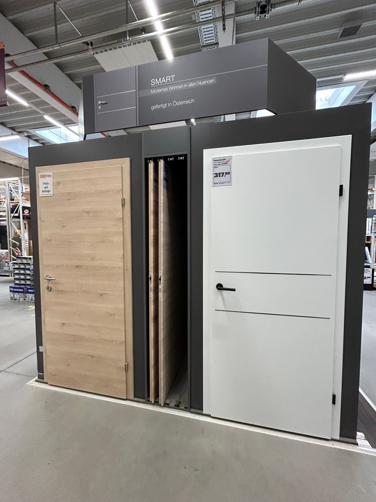 In einem Ausstellungsraum werden zwei Türen gezeigt: eine Holztür und eine weiße Tür. Die Holztür ist offen und zeigt einen schwarzen Griff. Die weiße Tür ist geschlossen. Ein Schild darüber lautet 'SMART' und 'Modernes Wohnen in allen Nuancen'. Ein Preisschild an der weißen Tür zeigt 317,90.