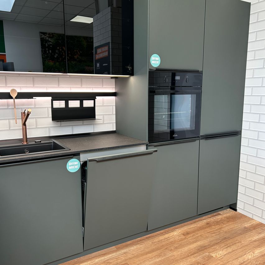 Eine moderne Küche mit schwarzem Backofen, grauen Schränken, einer Spüle und Holzboden. Ein Löffel und Wasserhahn sind über der Spüle, und ein Aufkleber sagt 'OFFRE MICHI'.