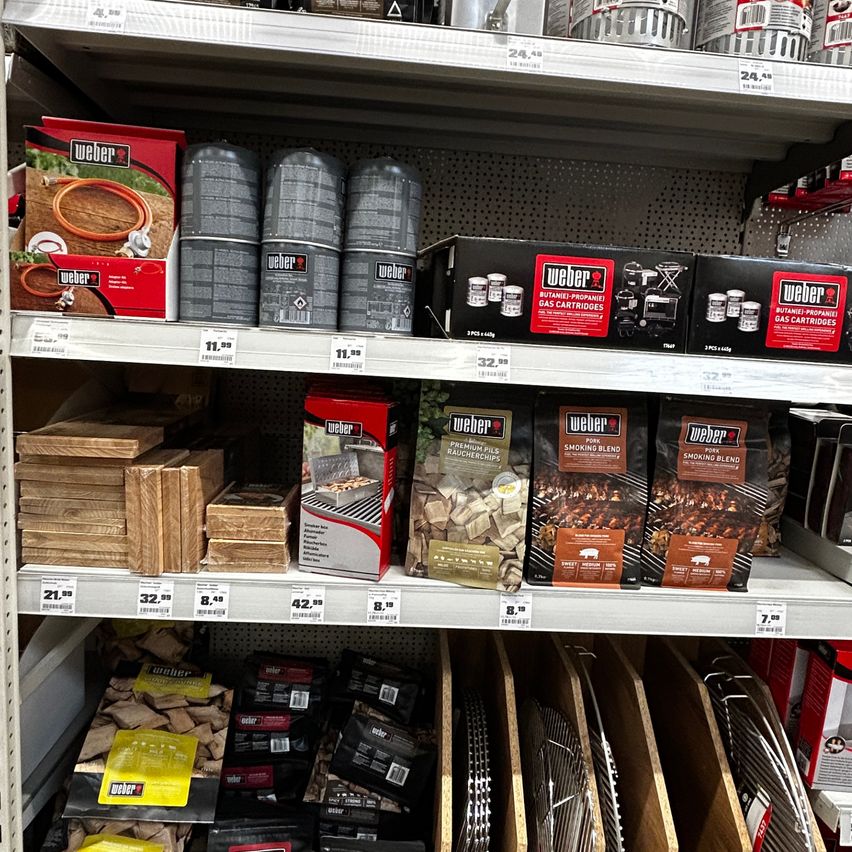 Ein Ladenregal zeigt verschiedene Weber-Grillprodukte, einschließlich Propangaskartuschen, Holzkohle und Räuchermischungen. Die Produkte sind ordentlich angeordnet, mit sichtbaren Preisschildern.