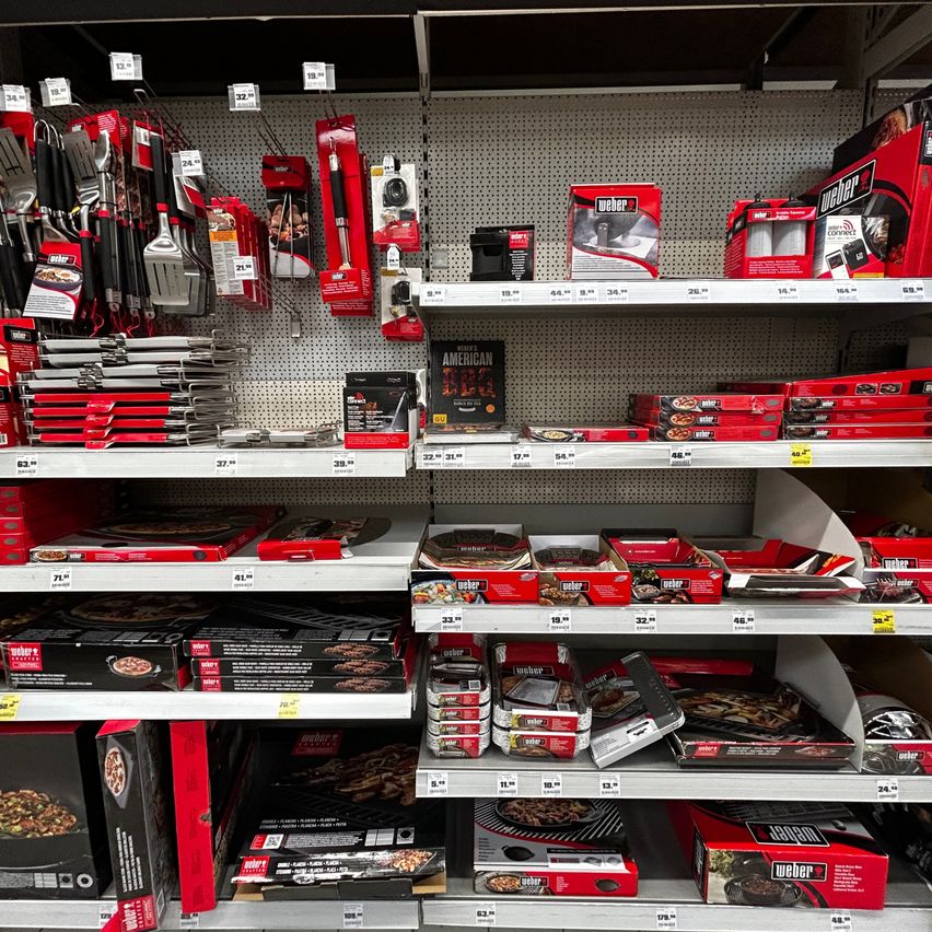 Ein Ladengang mit verschiedenen Grill- und Barbecue-Produkten, einschließlich Utensilien, Grillzubehör und verpackten Lebensmitteln. Die Regale sind mit roter und schwarzer Verpackung gefüllt, und Preisschilder sind sichtbar.