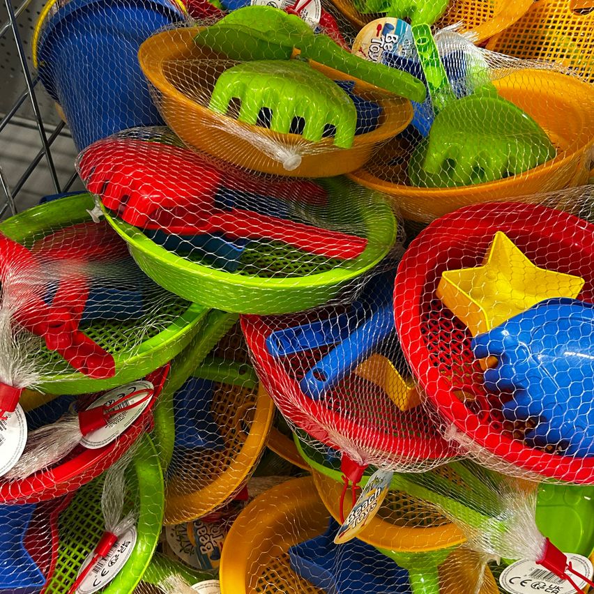 Eine Vielzahl von bunten Strandspielzeugen, einschließlich Schaufeln, Eimern und Netzen, sind in einem Geschäft ausgestellt. Jedes Spielzeug ist einzeln in Plastik verpackt und in einem Netzbeutel platziert.