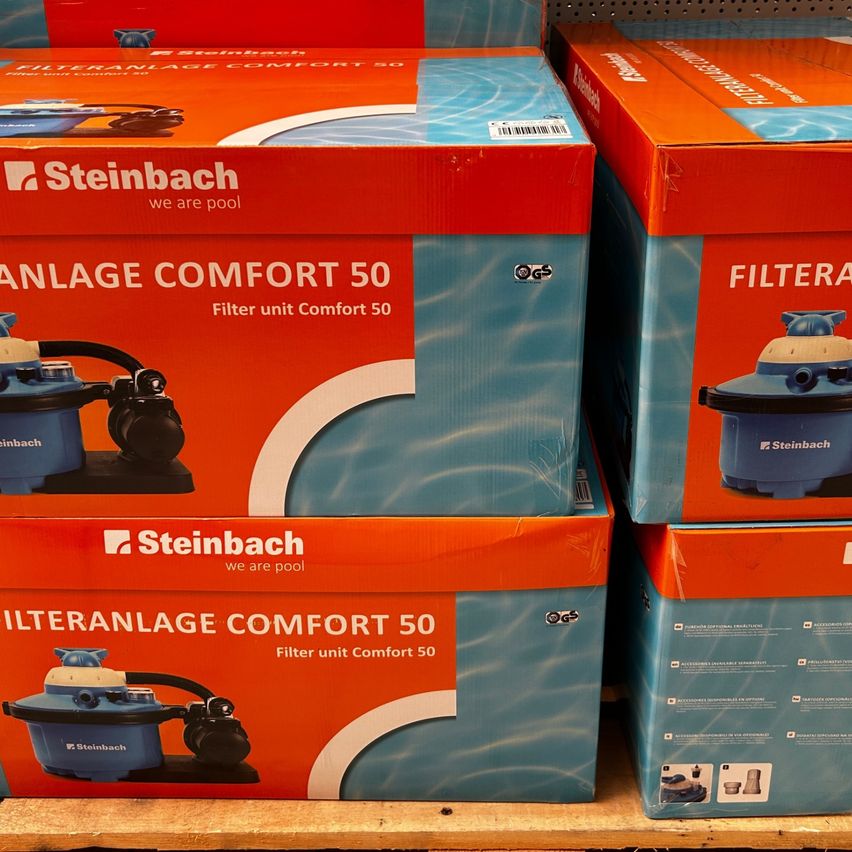 Gestapelte Kartons von Steinbach Filter Unit Comfort 50, auf einer Holzoberfläche ausgestellt. Die Kartons sind orange und blau mit einer Poolfilter-Illustration.