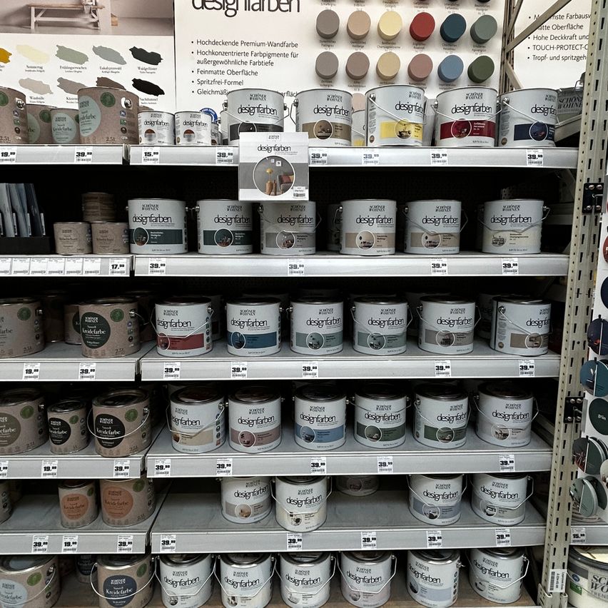 Ein Ladenregal gefüllt mit verschiedenen Farbdosen, die mit 'designfarben' beschriftet sind und eine Auswahl an Farben präsentieren. Die Dosen sind ordentlich in Reihen angeordnet, wobei einige eine kleine Musterkarte aufweisen.