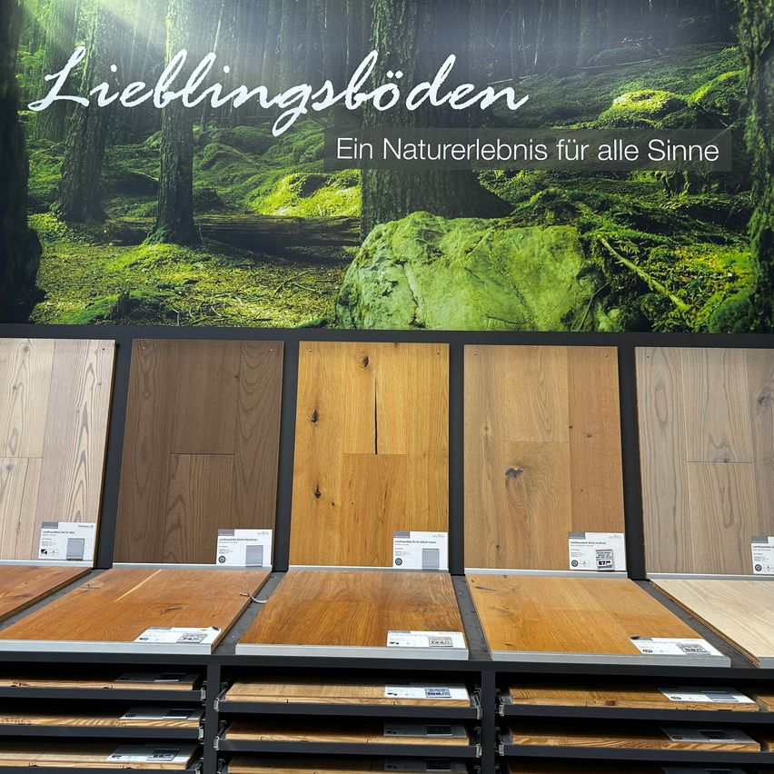 Eine Ausstellung verschiedener Holzbodenoptionen in einem Ausstellungsraum mit einem Waldhintergrund und einem Schild mit der Aufschrift 'Lieblingsboden'.