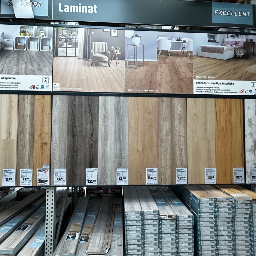 Ein Ladendisplay mit verschiedenen Holzlaminat-Bodenbelagsoptionen, mit Preisschildern und einem Schild, das 'Laminat Excellent' liest. Das Display enthält Bilder verschiedener Raumumgebungen.