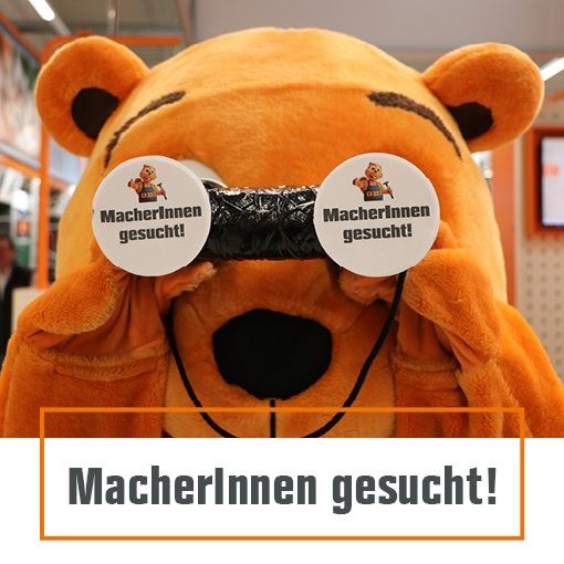 Ein Plüschbär-Maskottchen mit Fernglas und einem Schild, auf dem 'MacherInnen gesucht!' steht. Der Maskottchen hält das Fernglas mit beiden Pfoten und bedeckt die Augen. Das Schild erscheint zweimal, einmal auf jeder Seite des Fernglases.