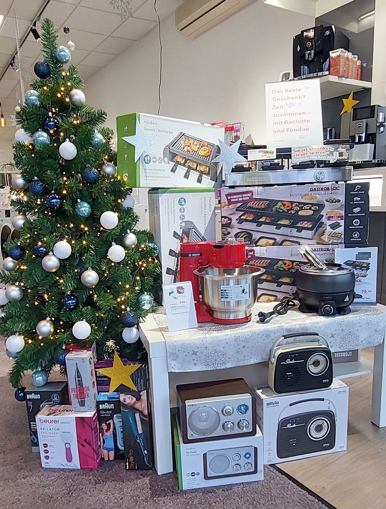 Ein Weihnachtsbaum mit Ornamenten steht neben einem Tisch, auf dem verschiedene Küchengeräte stehen, darunter ein roter Mixer, ein schwarzer Reiskocher und ein silbernes Gerät. Zusätzliche Kartons sind um den Baum und auf dem Boden verstreut. Ein Sternenschild und ein weißes Schild mit Text hängen darüber.