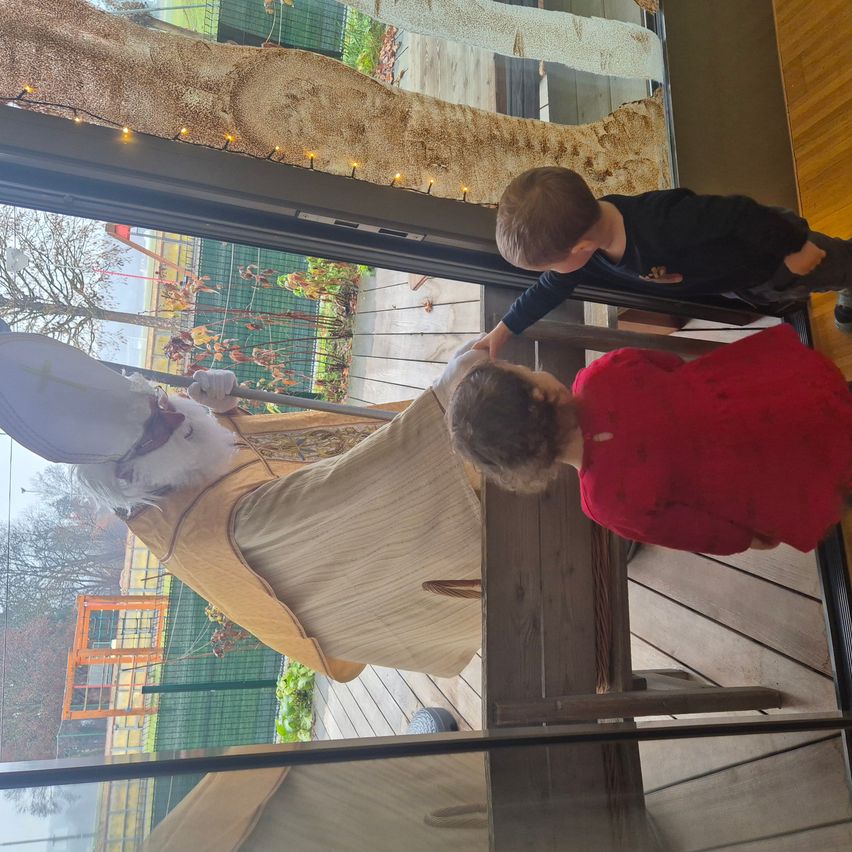 Zwei Kinder spielen mit einer großen weißen Statue in einem Gebäude. Die Statue hat ein weißes Tuch über ihrem Körper, und die Kinder berühren sie. Außerhalb des Gebäudes gibt es einen Baum mit Blättern und einen Zaun.