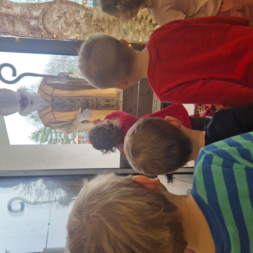 Eine Gruppe von Kindern beobachtet einen als Priester verkleideten Menschen durch ein Glasfenster. Die Person trägt eine weiße Robe und hält einen Stab.