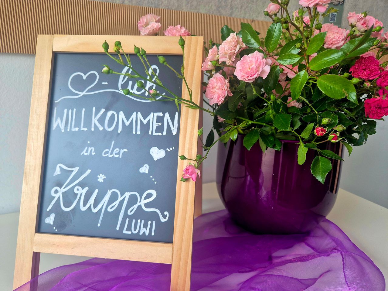 Ein Holzschild mit einem Strauß rosa Blumen und einem lila Blumentopf auf einem lila Tuch. Das Schild sagt 'Willkommen in der Krüppe LULU'.