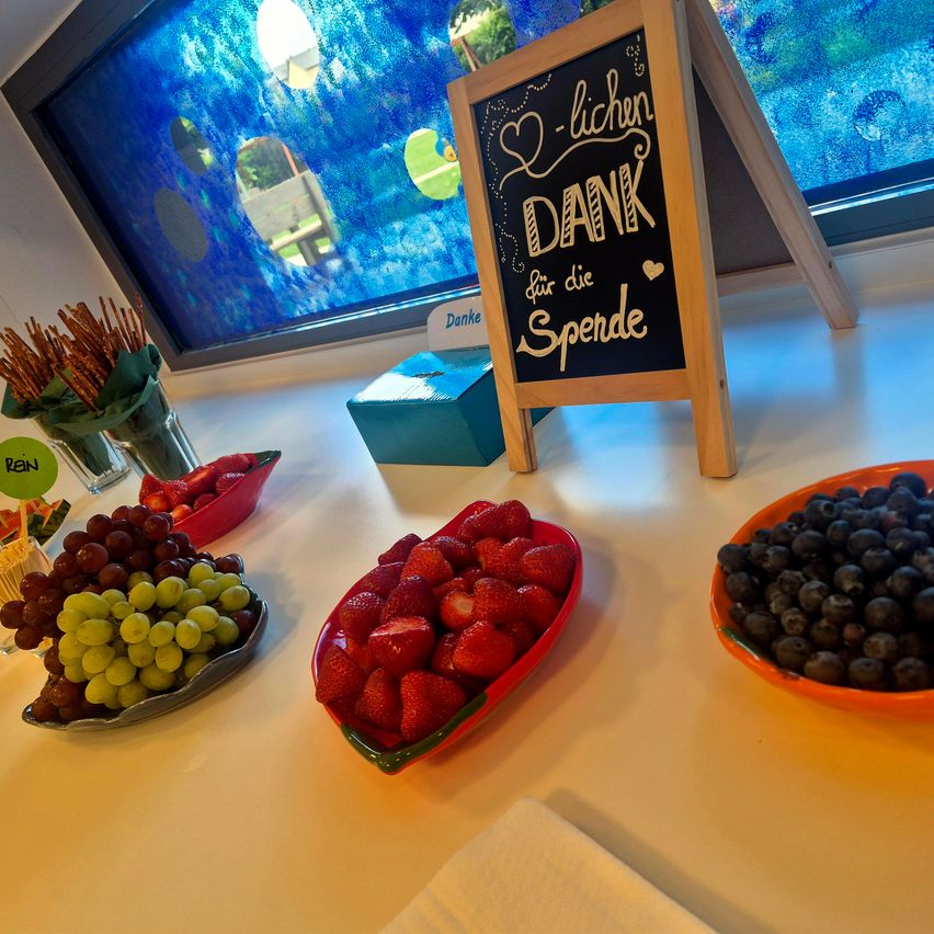 Ein Kreidetafel mit dem Text 'Ich-lichen DANK für die Spende' steht neben Schalen mit Erdbeeren, Trauben und Blaubeeren. Eine klare Vase mit Brezeln und eine blaue Box sind ebenfalls vorhanden.