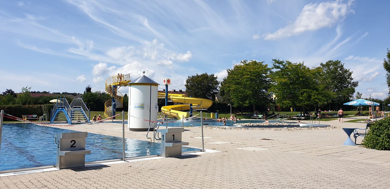 Ein Schwimmbad mit einer gelben Rutsche und einer Sprungplattform, umgeben von einem Zaun, unter einem blauen Himmel mit Wolken.