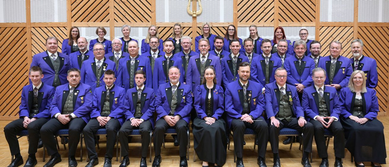 Ein Chor in passenden blauen Anzügen und schwarzen Krawatten posiert für ein Gruppenfoto in einem Raum mit Holzwänden und einem goldenen Symbol darüber.