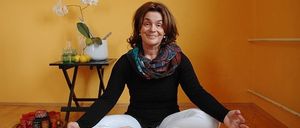 Eine Frau sitzt mit gekreuzten Beinen auf einem Holzboden, trägt ein schwarzes Shirt und weiße Hosen und lächelt. Hinter ihr befindet sich eine gelbe Wand mit einem kleinen Tisch, auf dem eine weiße Vase mit Blumen und ein Teller mit roten und gelben Objekten stehen.