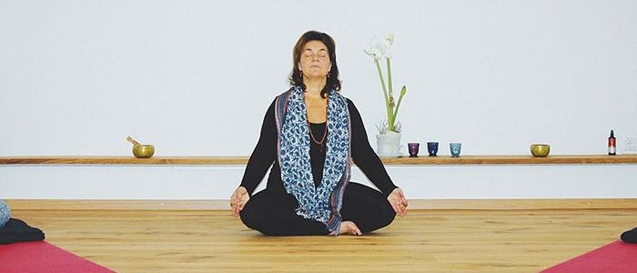 Eine Frau praktiziert Yoga auf dem Boden. Sie trägt einen Schal und befindet sich in der Lotusposition. Hinter ihr steht eine weiße Wand, auf deren Regal sich eine Vase mit weißen Blumen und einigen Schalen befinden.