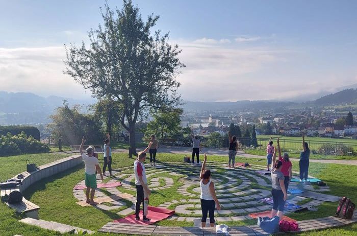 Eine Gruppe von Menschen praktiziert Yoga auf einem kreisförmigen Pfad in einem Park. Sie sind von einem Rasengebiet mit einem Baum und einer entfernten Aussicht auf die Stadt umgeben.
