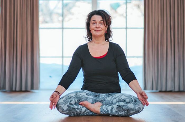 Eine Frau sitzt in einer Lotusposition auf einem Holzboden, lächelt und hat ihre Hände in einer Gebetsposition.