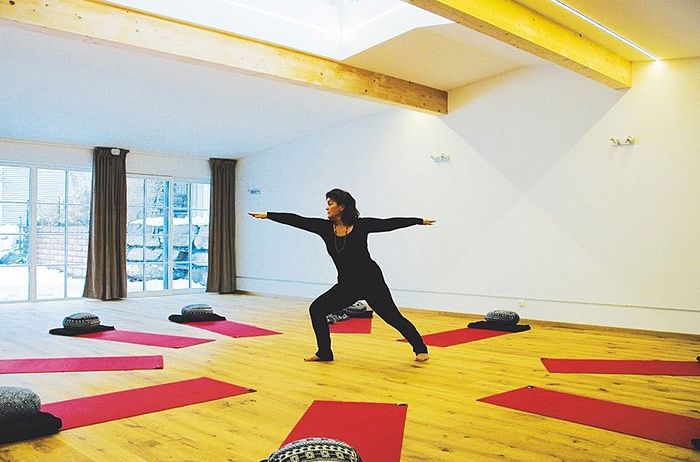 Eine Frau praktiziert Yoga in einem geräumigen, hellen Studio. Sie steht mit ausgestreckten Armen, umgeben von roten Yogamatten und Kissen. Holzbalken und Glasfenster sind sichtbar.