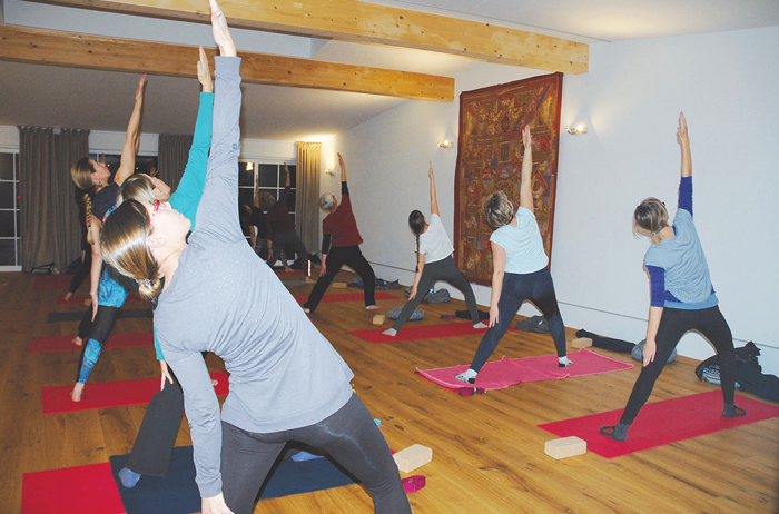 Eine Gruppe von Menschen praktiziert Yoga in einem Studio mit Holzböden und weißen Wänden. Sie tragen alle Yoga-Kleidung und verwenden rote Yoga-Matten. Einige befinden sich in Posen, während andere zwischen den Posen wechseln.