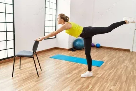 Eine Frau in Trainingskleidung führt eine Yoga-Pose durch, die sie mit einem Stuhl ausbalanciert, in einem Fitnessstudio mit Holzböden.
