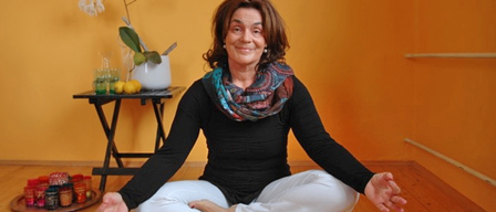 Eine Frau sitzt auf dem Boden, lächelt und hat ihre Hände in einer Meditationshaltung. Sie trägt ein schwarzes Shirt und weiße Hose. Hinter ihr steht ein Tisch mit einer weißen Vase mit weißen Blumen und gelben Zitronen. Ein Regal befindet sich an der Wand.