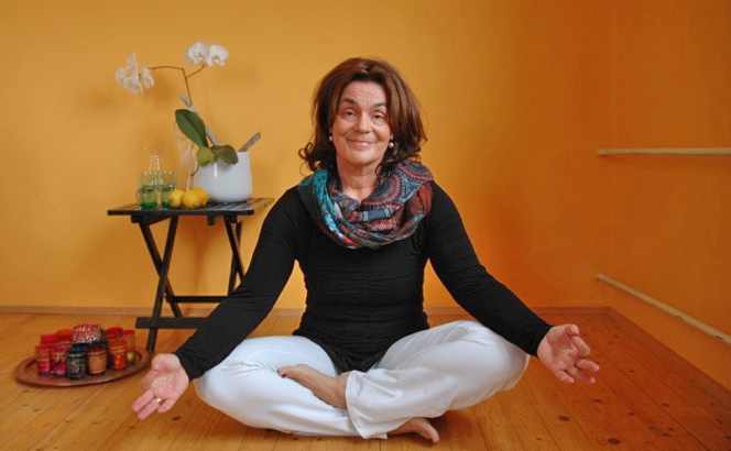 Eine Frau sitzt auf dem Boden, lächelt und hat ihre Hände in einer Meditationshaltung. Sie trägt ein schwarzes Shirt und weiße Hose. Hinter ihr steht ein Tisch mit einer weißen Vase mit weißen Blumen und gelben Zitronen. Ein Regal befindet sich an der Wand.