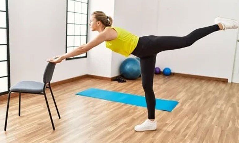Eine Frau führt eine Beinübung mit einem Stuhl in einem Fitnessstudio durch und hält sich dafür an ihm fest. Sie trägt ein gelbes Tanktop und schwarze Leggings. Yogamatten und Übungsbälle sind in der Nähe.