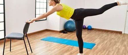 Eine Frau führt eine Beinübung mit einem Stuhl in einem Fitnessstudio durch und hält sich dafür an ihm fest. Sie trägt ein gelbes Tanktop und schwarze Leggings. Yogamatten und Übungsbälle sind in der Nähe.