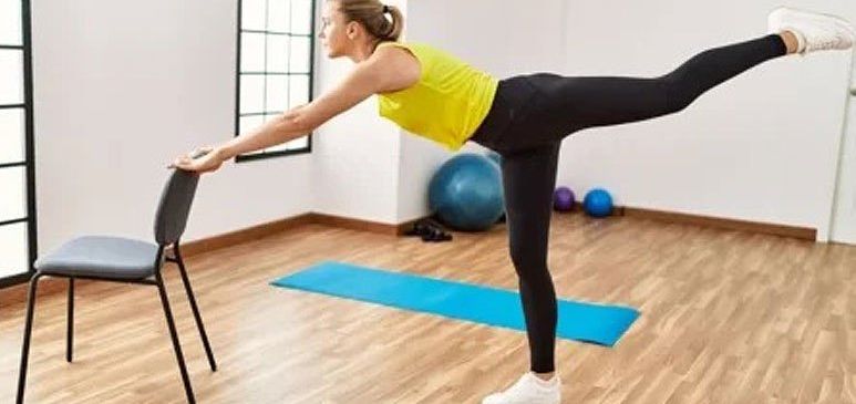 Eine Frau führt eine Beinübung mit einem Stuhl in einem Fitnessstudio durch und hält sich dafür an ihm fest. Sie trägt ein gelbes Tanktop und schwarze Leggings. Yogamatten und Übungsbälle sind in der Nähe.