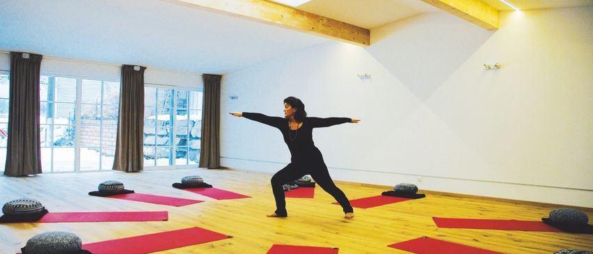 Eine Frau in einem Studio praktiziert Yoga auf einem Holzboden mit roten Matten und Kissen. Der Raum hat Holzbalken und weiße Wände.