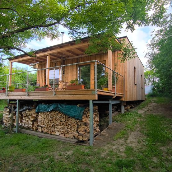 Ein Holzhaus mit Balkon und einem kleinen Holzstapel darunter. Das Haus ist von üppiger Grünfläche und Bäumen umgeben.