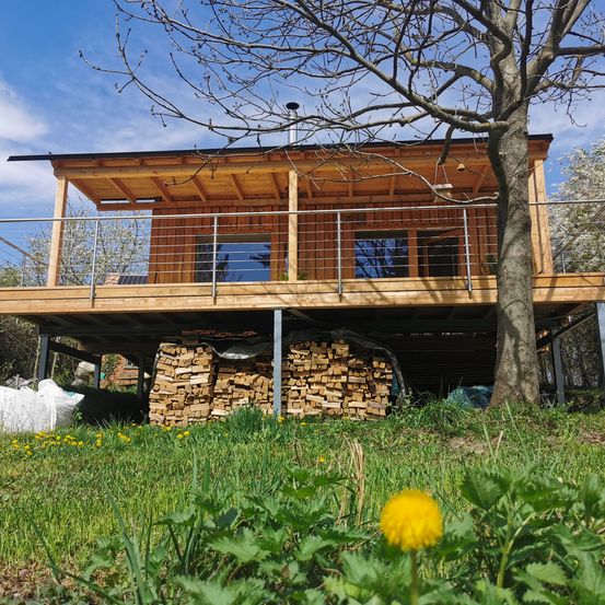 Ein Holzhaus mit Balkon und Schornstein steht auf einer Holzplattform, umgeben von einem Holzstapel und grünem Gras.