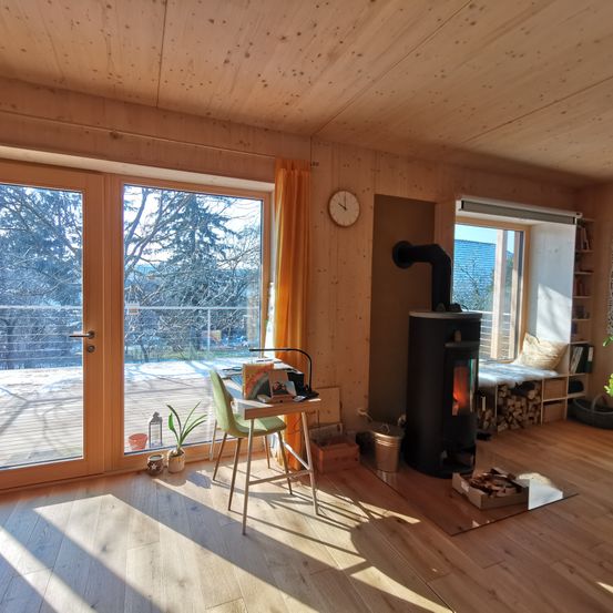 Ein gemütlicher Raum mit Holzwänden und -böden. Es gibt einen Schreibtisch mit Lampe und Büchern. Eine Holzheizung mit Feuer steht in der Ecke. Ein Balkon mit Geländer bietet Blick auf eine verschneite Landschaft.