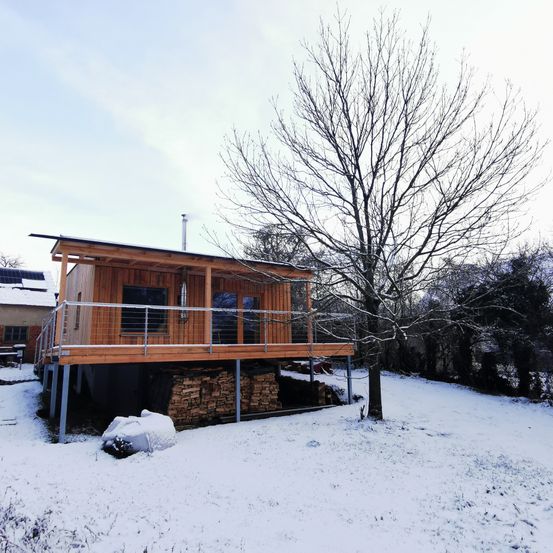 Ein kleines Holzhaus mit Balkon und Schornstein ist von Schnee umgeben. In der Nähe befinden sich Holzstapel und ein kahler Baum.