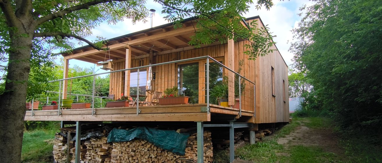 Ein Holzhaus mit Balkon steht auf einem Hügel, mit einem Holzstapel darunter. Bäume und Pflanzen umgeben das Haus.