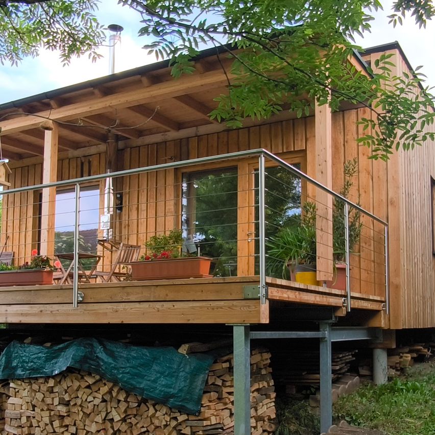 Ein Holzhaus mit Balkon steht auf einem Hügel, mit einem Holzstapel darunter. Bäume und Pflanzen umgeben das Haus.