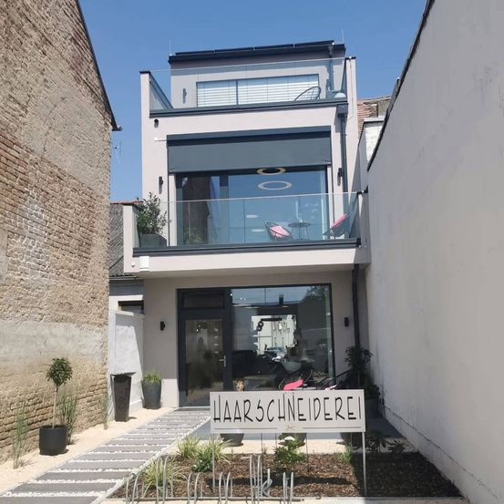 Ein modernes Haus mit Balkon, Glasfenstern und einem Schild, das 'Haarschneiderei' besagt. Das Haus ist von Pflanzen umgeben und hat eine Ziegelmauer auf der linken Seite.