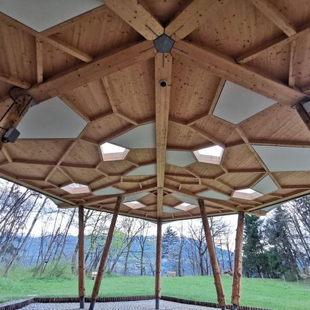 Ein hölzernes Pavillon mit einer sechseckigen Dachkonstruktion, die dreieckige Oberlichter und Holzbalken aufweist. Es ist von einem Grasfeld mit Bäumen und Bergen im Hintergrund umgeben.