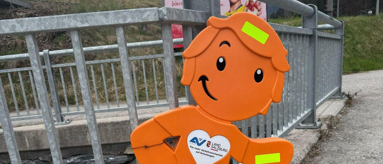 Eine orangefarbene Figur eines lächelnden Jungen steht auf einem Beton-Gehweg neben einem Metallgeländer. Der Junge hat ein weißes Herz auf der Brust und gelbe Reflektoren an Armen und Beinen.