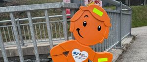 Eine orangefarbene Figur eines lächelnden Jungen steht auf einem Beton-Gehweg neben einem Metallgeländer. Der Junge hat ein weißes Herz auf der Brust und gelbe Reflektoren an Armen und Beinen.